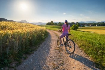 Woman on mountain bike on gravel road at sunset in summer. Colorful landscape with sporty girl riding bicycle, 
blooming fields, meadows with yellow and green grass, dirt road, sky. Sport and travel