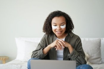 Modern mixed race girl with under eye patches on face working at laptop in bedroom. Biracial young lady with under-eye patch watching series on computer, sitting on bed. Skincare beauty routine.. Mixed race girl with under eye patches on face working at laptop in bedroom. Skincare beauty routine