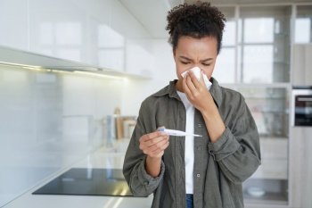 Sick mixed race girl blowing nose checks temperature using electronic thermometer at home. Young biracial teen suffer from influenza or cold, wiping runny nose with napkin. Flu fever, covid pandemic.. Sick mixed race girl blowing nose checks temperature using electronic thermometer at home. Flu fever