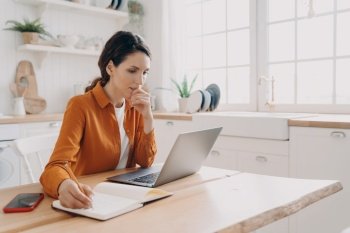 Focused woman works at laptop online from home, writing notes, sitting at table. Pensive housewife plans purchases, writes to-do list for week, sitting in kitchen. Distant job, domestic life.. Female housewife freelancer works at laptop, plans purchases, notes, sitting in kitchen at home
