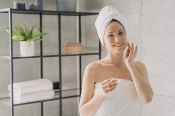 Beautiful hispanic woman wearing towel on head using cotton pad, cleanses facial skin. Smiling female removing makeup standing in bathroom. Skincare cleansing cosmetics advertising.. Girl cleanses face skin by cotton pad standing in bathroom. Skincare cleansing cosmetics advertising