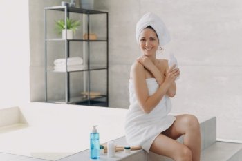 Pretty woman holding cosmetic bottle sitting on bathtub in modern bathroom. Smiling latina female wrapped in towel advertising cosmetics product for body care. Skincare, spa treatment procedure.. Pretty woman advertising cosmetics for body care in bathroom. Skincare treatment, spa procedure