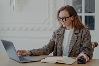 Elegant happy mid adult businesswoman is doing research, browsing internet at home and smiling. Emotional european lady in jacket is freelancer, entrepreneur or accountant. Remote work at the desk.. Elegant businesswoman browsing internet at home and smiling. Remote work at the desk.