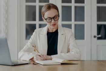Elegant concentrated businesswoman in formal wear. Glamorous mid adult business lady is working from home and taking notes. Attractive european woman is a boss or entrepreneur. Remote work concept.. Elegant concentrated businesswoman in formal wear is working from home and taking notes.