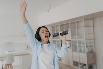 Energetic female wearing wireless headphones sing karaoke, using TV remote control like a microphone, rests at home. Excited woman singing a song, raised her arm, enjoying music sound.. Energetic female in headphones sing karaoke, using TV remote control like a mic, rests at home