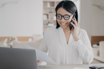 Smiling businesswoman talking on phone, answering call, works with documents, sitting at laptop. Happy female manager in glasses consulting client or enjoying corporate mobile conversation in office.. Smiling businesswoman talking on phone, answering call, works with documents, sitting at laptop
