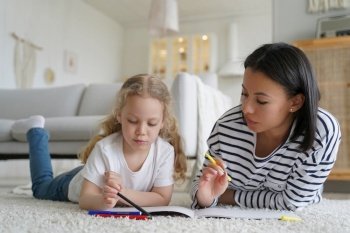 Mom or babysitter and adopted little daughter drawing, lying on warm floor at home. Mother teaches kid girl to draw in sketchbook with colorful pencils. Educational pastime, children’s creativity.. Mom teaches kid daughter to draw by colorful pencils, lying on floor at home. Children’s creativity