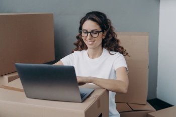 Smiling hispanic female wearing glasses working at laptop surrounded with cardboard boxes, preparing customers orders, parcels of online store for sending. Delivery service, e-commerce concept.. Girl works at laptop preparing parcels of online store for sending. Delivery service, ecommerce