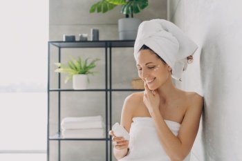 Smiling woman holds white jar with cleansing foam or moisturizing serum for facial treatment, female with towel on head using rejuvenating cream in bathroom at home. Skincare daily routine.. Smiling woman using moisturizing cream for facial treatment in bathroom. Skincare daily routine