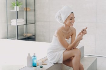 Smiling young woman wrapped in towel reading ingredients list on cosmetics sample sitting on bathtub in bathroom. Skincare treatment, spa procedure, natural cosmetic products advertising.. Smiling woman wrapped in towel holds cosmetics sample sitting in bathroom. Skincare treatment, spa