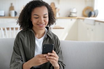 Happy mixed race teen girl holding smartphone uses mobile apps, shopping online, enjoying web surfing on internet. Smiling young lady with afro hair communicating in social networks at home.. Happy mixed race teen girl holding smartphone uses mobile apps, chatting or shopping online at home