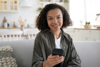 Cheerful happy mixed race teen girl holding smartphone with modern mobile apps, looking at camera, sitting on sofa. Smiling young lady sits on couch using online app on phone indoors.. Smiling mixed race girl holding smartphone with modern apps, looking at camera, sits on sofa at home
