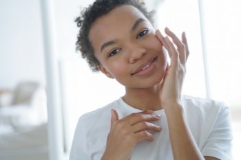 Smiling pretty african american young girl touching under eye skin, looking in the mirror. Beautiful lady enjoying skincare, applying moisturizing facial cream. Natural beauty, cosmetics advertising.. Smiling pretty african american young girl enjoying skincare. Natural beauty, cosmetics advertising