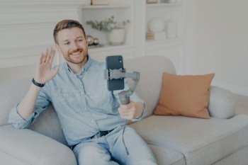 Young man enjoying video chat with friends , looking at phone screen and waving with hand, cheerful bearded man holding smartphone with gimbal stabilizer handheld while sitting on sofa in living room. Young man enjoying video chat with family on cellphone while sitting on sofa in living room