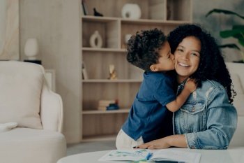 Portrait of happy afro american family at home, cute boy in blue polo tshirt expressing his love by kissing in cheek and hugging mother who sits on floor in living room, enjoying time together. Portrait of afro american cute boy in blue polo tshirt hugging mother and kissing her in cheek