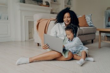 Young positive mixed race mother and cute boy son having video call with relatives, father or grandparents, sitting with laptop on floor and smiling, child wearing headset and pointing at screen. Young positive mixed race mother and cute boy son having video call on laptop