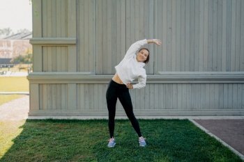 Sporty lifestyle wellness and weight loss concept. Active woman in sportwear trains outdoors leans left stands on green grass warms up before workout has active life enjoys regular workout in open air