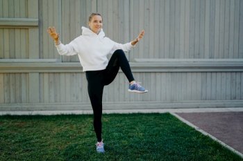 Energetic sportwoman has workout routine poses on one legs demonstrates her stamina warms up before jogging dressed in hoodie leggings and trainers trains outdoors. Sport and motivation concept