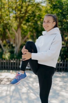 Vertical shot of energetic woman leads active lifestyle keeps knee raised exercises outdoor dressed in active wear lifts leg smiles pleasantly listens audio track in wireless earphones. Workout