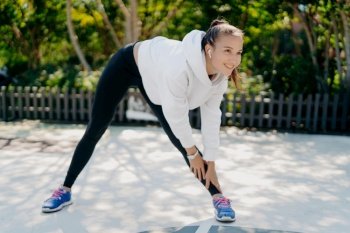 Photo of active pleased woman smiles gladfully concentrated into distance leans to leg dressed in sportswear trains outdoors being motivated listens music in wireless earphones does aerobic exercises