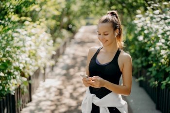 Sport as style of life. Horizontal shot of healthy slim sporty woman focused at smartphone checks information and chooses song from playlist dressed in sportwear poses outdoor breathes fresh air