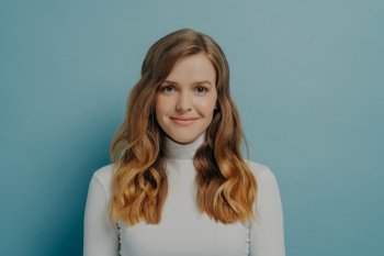 Half-length portrait of beautiful dreamy millennial woman with wavy hair in white turtleneck looking at camera with gentle smile isolated over pastel blue studio wall background with copy space. Beautiful dreamy young woman with perfect wavy styled hair in white turtleneck smiling at camera