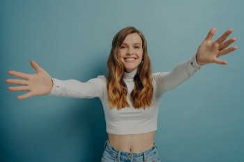 Half-length photo of overjoyed cheerful young woman standing in front of camera smiling with arms wide open outstretched to embrace hug someone desirable and beloved isolated on blue background. Overjoyed cheerful young woman with arms wide open outstretched to embrace hug someone