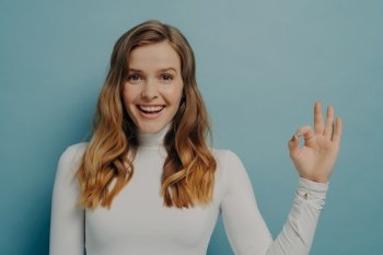 Half-length image of beautiful excited joyful young lady with dyed light-brown hair gesturing ok symbol with fingers, looking at camera with beaming smile isolated over blue studio wall background. Beautiful excited joyful young lady with dyed light-brown hair gesturing ok symbol with fingers