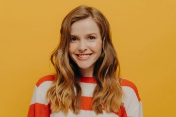Keep smiling. Studio portrait of cute teen girl looking at camera with toothy smile, sending positive energy to someone while posing isolated over yellow background in striped red and white sweater. Happy teen girl sending positive energy to someone while standing isolated over yellow background