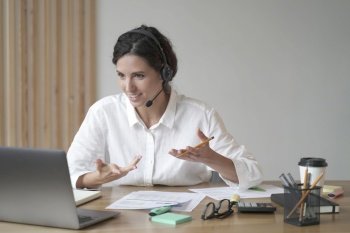 Smiling european woman online tutor sitting at desk in headset and talking by video call on laptop computer while working remotely in home office. Distance education and e-learning concept. Smiling woman online tutor sitting at desk in headset and talking by video call on laptop computer