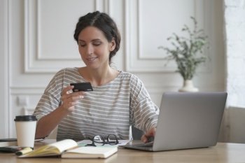 Online shopping. Young smiling italian woman in casual clothes looking at credit card and typing on laptop, paying online, buying goods or services in internet store while sitting at table at home. Young smiling italian woman in casual clothes looking at credit card and typing on laptop