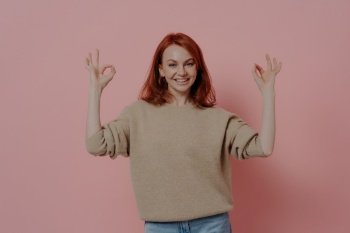 Half length shot of pretty ginger woman showing OK sign with both hands, saying that everything is fine, dressed in casual outfit, satisfied red-haired model posing isolated over pink background. Half length shot of pretty ginger woman showing OK sign with both hands, isolated on pink