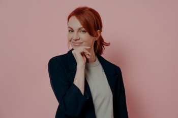 Successful europian lady with red hair isolated over pink backround, touching chin with hand and looking at camera with confident smile, happy red-hahaired female enterpreneur or business woman. Successful europian lady with red hair isolated over pink backround, touching chin with hand