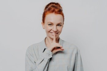 Sign of silence. Headshot portrait of cheerful young ginger 30s woman showing hush gesture, keeping finger on lips while posing isolated on grey studio background. Privacy and silence concept. Cheerful young ginger 30s woman showing hush gesture while posing isolated on grey studio background