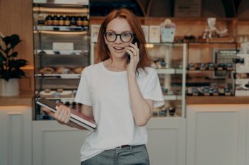 Busy female bakery shop owner has conversation on mobile phone holds notepads poses indoor wears spectacles casual white t shirt looks into distance. People technology and occupation concept. Busy female bakery shop owner has conversation on mobile phone