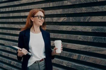 Glad redhead young woman enjoys life has successful day good mood drinks takeaway coffee holds digital tablet and notepad looks happily into distance wears formal clothes poses outdoor near wall. Glad redhead young woman drinks takeaway coffee holds digital tablet and notepad