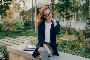 Tired redhead woman takes rest in park suffers from neck pain sits on bench with takeaway coffee notebook digital device dressed formally poses outdoor. Businesswoman feels fatigue after busy day. Tired redhead woman takes rest in park suffers from neck pain sits on bench