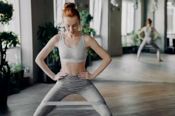 Slender sportive redhead lady performing exercises with fitness elastic band on legs, keeping hands on waist, looking down at toes in gym with large mirror behind. Healthy lifestyle and sport indoor. Slender sportive redhead lady performing sit ups with fitness elastic band