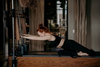 Side view of athletic young caucasian female stretching body muscles while exercising on cadillac reformer in modern fitness studio, wearing black sports clothes. Pilates training and sport indoor. Flexible ginger woman stretching arms on cadillac reformer