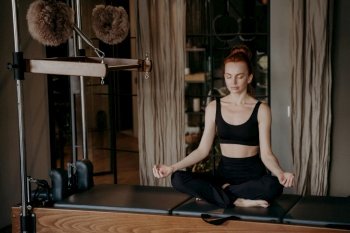 Peaceful female pilates instructor sitting in yoga lotus position on Cadillac reformer, trying to relax after intensive stretching exercises in pilates studio. Meditation and yoga concept. Young redhead woman in sportswear sitting in lotus position on trapeze table trying to relax