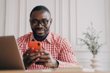 Cheerful smiling African ethnicity man office employee sitting at desk with open laptop holding smartphone chatting with friends while working remotely online from home. Freelance concept. Smiling African ethnicity man office worker sitting at desk holding smartphone chatting with friends