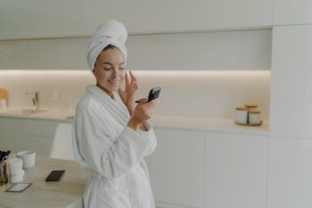Take care of yourself. Young happy woman in bathrobe looking with smile in compact mirror and applying skin care product on face while standing in modern kitchen and doing cosmetic procedures at home. Happy healthy woman in bathrobe applying skin care product on face after taking shower or bath at home