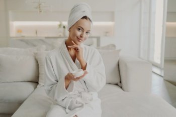 Portrait of young pretty woman with in white bathrobe and towel on head holding cosmetic cream, touching her soft healthy face skin and smiling at camera while relaxing on sofa in cozy apartment. Young beautiful woman holding cosmetic product and smiling while relaxing on sofa after taking shower at home
