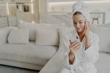 Young beautiful happy woman female beauty blogger wearing white bathrobe and towel on head applying face cream while creating video on smartphone for social media or vlog about daily skincare routine. Woman beauty blogger creating video on smartphone for her vlog about daily skincare routine