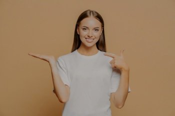 Half length shot of good looking brunette young woman points at raised palm pretends holding something advertises some item wears casual t shirt isolated over brown background. Promotion concept