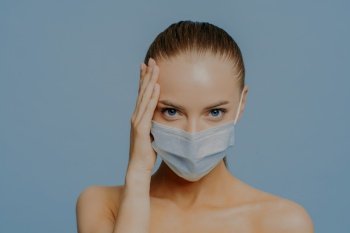 Covid 19 concept. Serious brunette European woman touches face looks confidently at camera has combed hair makeup wears medical protective mask during virus pandemic stands shirtless indoor.
