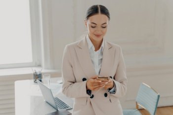 Happy beautiful female business consultant in formal wear using mobile phone, reading news or sending text message while standing indoor at modern light office, enjoying busy working day. Young businesswoman reading news or sending text message