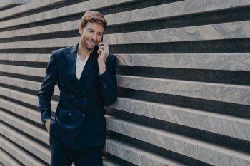 Half length shot of successful businessman calls via smartphone during work break talks with business partner dressed in formal clothes stands near marble wall copy space for your promotion.. Successful businessman calls via smartphone during work break talks with business partner