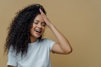 Horizontal shot of happy positive Afro American woman has fun, keeps hand on forehead, giggles happily, closes eyes, has bushy curly hair, wears casual white t shirt, isolated on beige background.