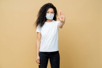 Say no to spreading disease. Serious looking Afro American woman makes stop gesture, wears medical mask, prevents virus Covid-19, stands indoor, isolated. Stop infectious disease immediately